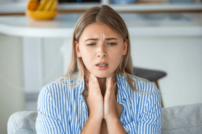tonsil-stone-symptoms-throat-pain A young woman with blonde hair, wearing a blue and white striped shirt, sits on a couch, holding her throat with both hands. She has a pained or uncomfortable expression, indicating throat discomfort. The background shows a kitchen with a fruit bowl on the counter.